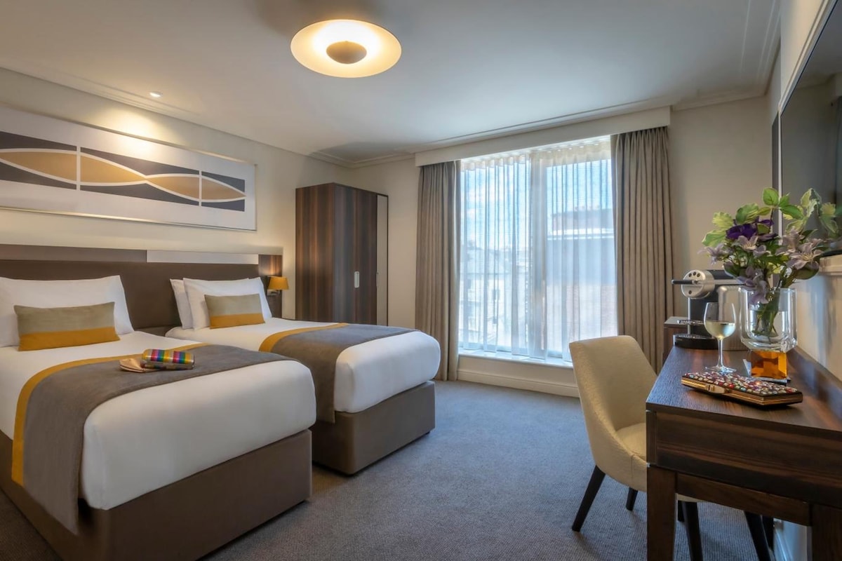 A hotel room with two beds, a desk, a chair, a coffee maker, and a window with curtains.