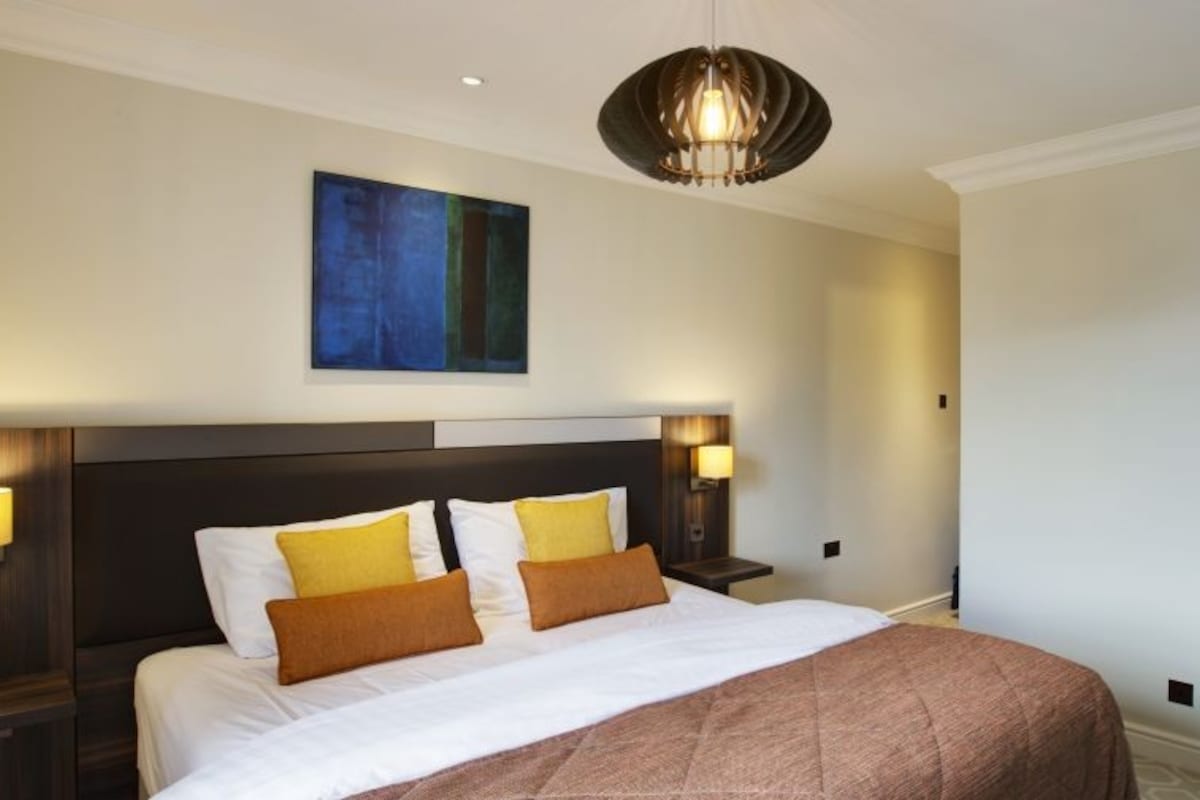 A hotel room with a large bed, two bedside lamps, a modern headboard, a painting on the wall, and a ceiling light fixture.