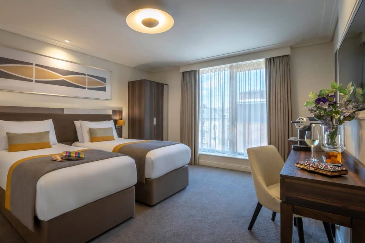 Habitación de hotel con dos camas, un escritorio, una silla, una cafetera y un ventanal con cortinas.