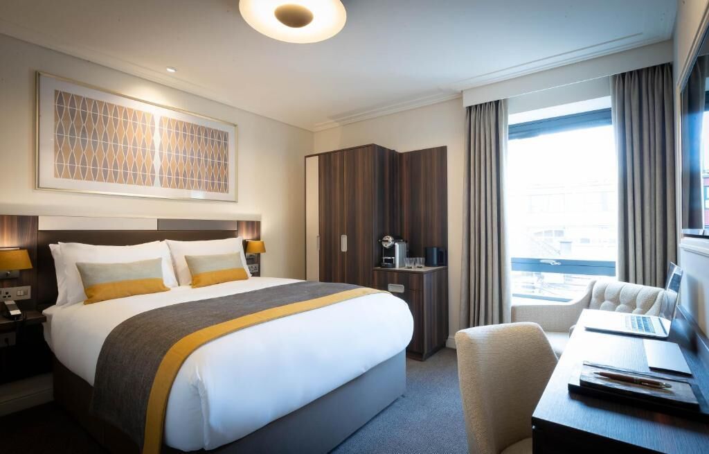 A hotel room with a large bed, a desk with a laptop, a wardrobe, and a view of the city.