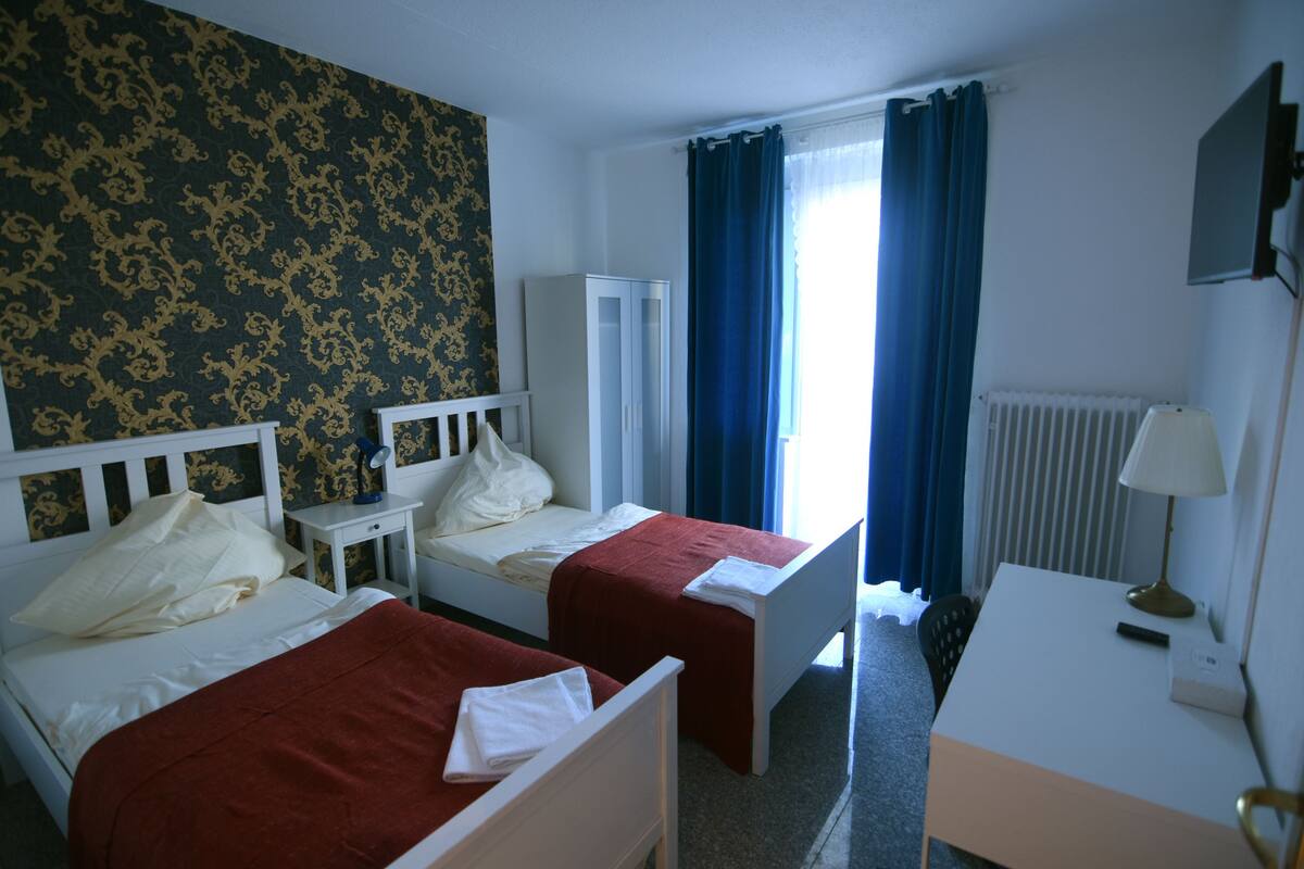 A hotel room with two single beds, a small table, a chair, a desk, a lamp, a mirror, and a radiator.