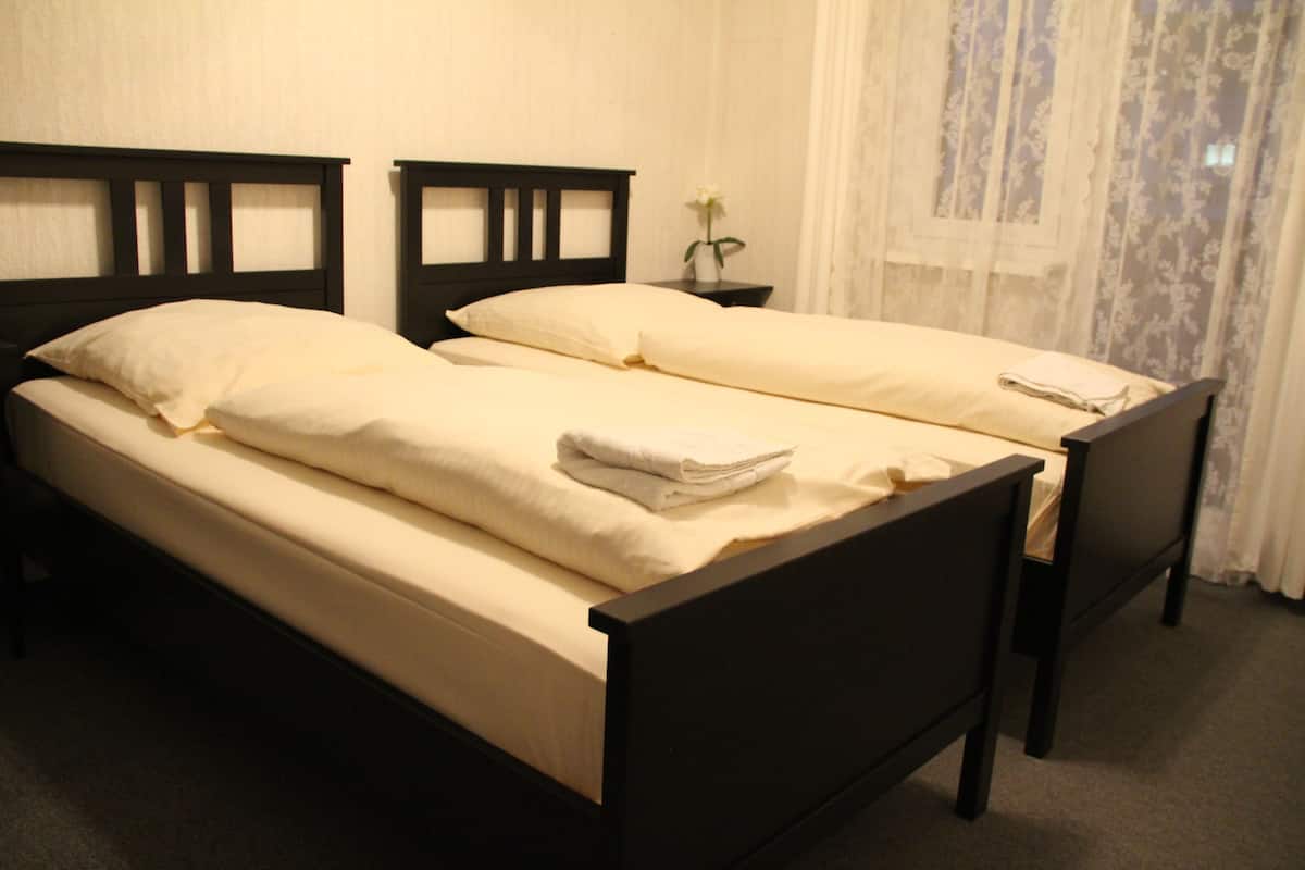 A double bed with a wooden headboard, a nightstand with a vase of flowers, and a window with sheer curtains.