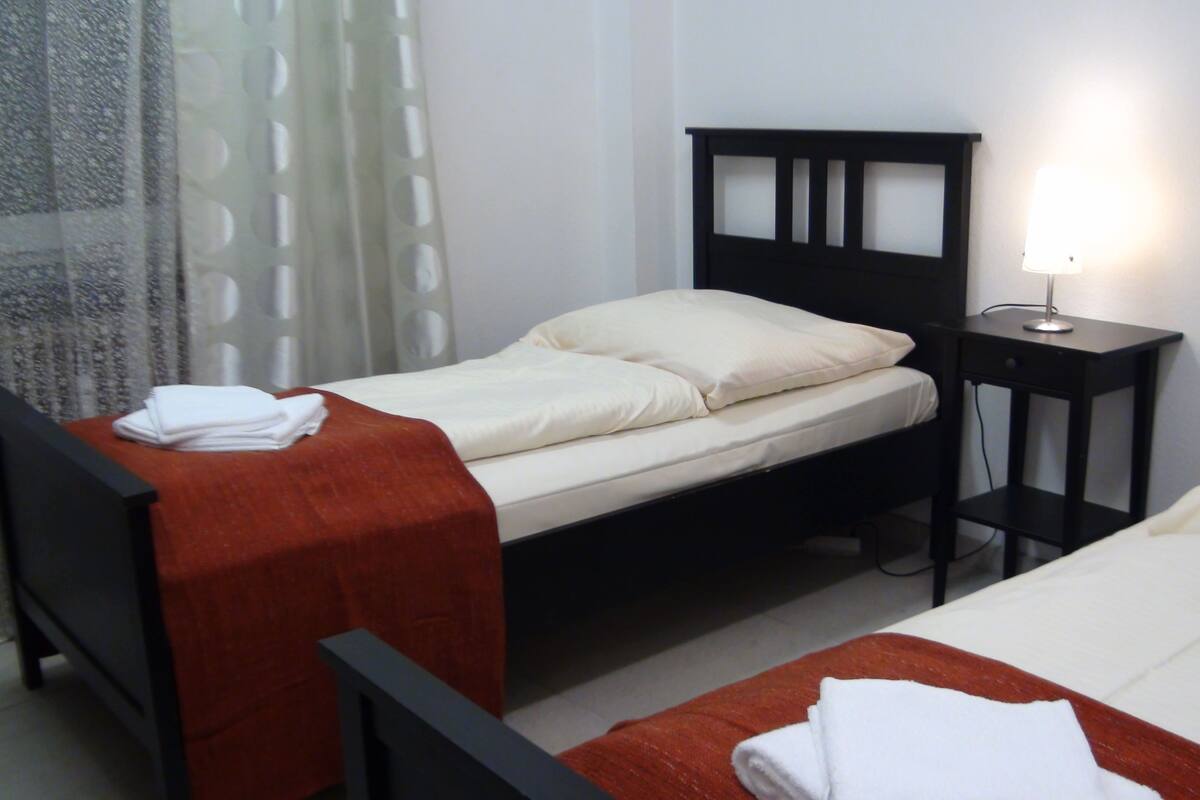 A twin room with two beds, a bedside table with a lamp, and a window with curtains.