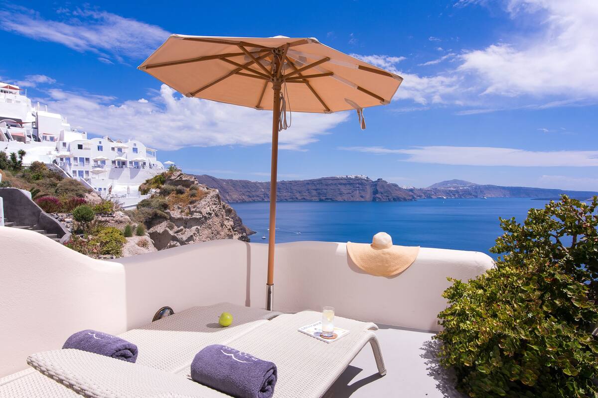 A sun lounger with a beige umbrella, a hat, and towels, overlooking a coastal view with white buildings and blue sea.