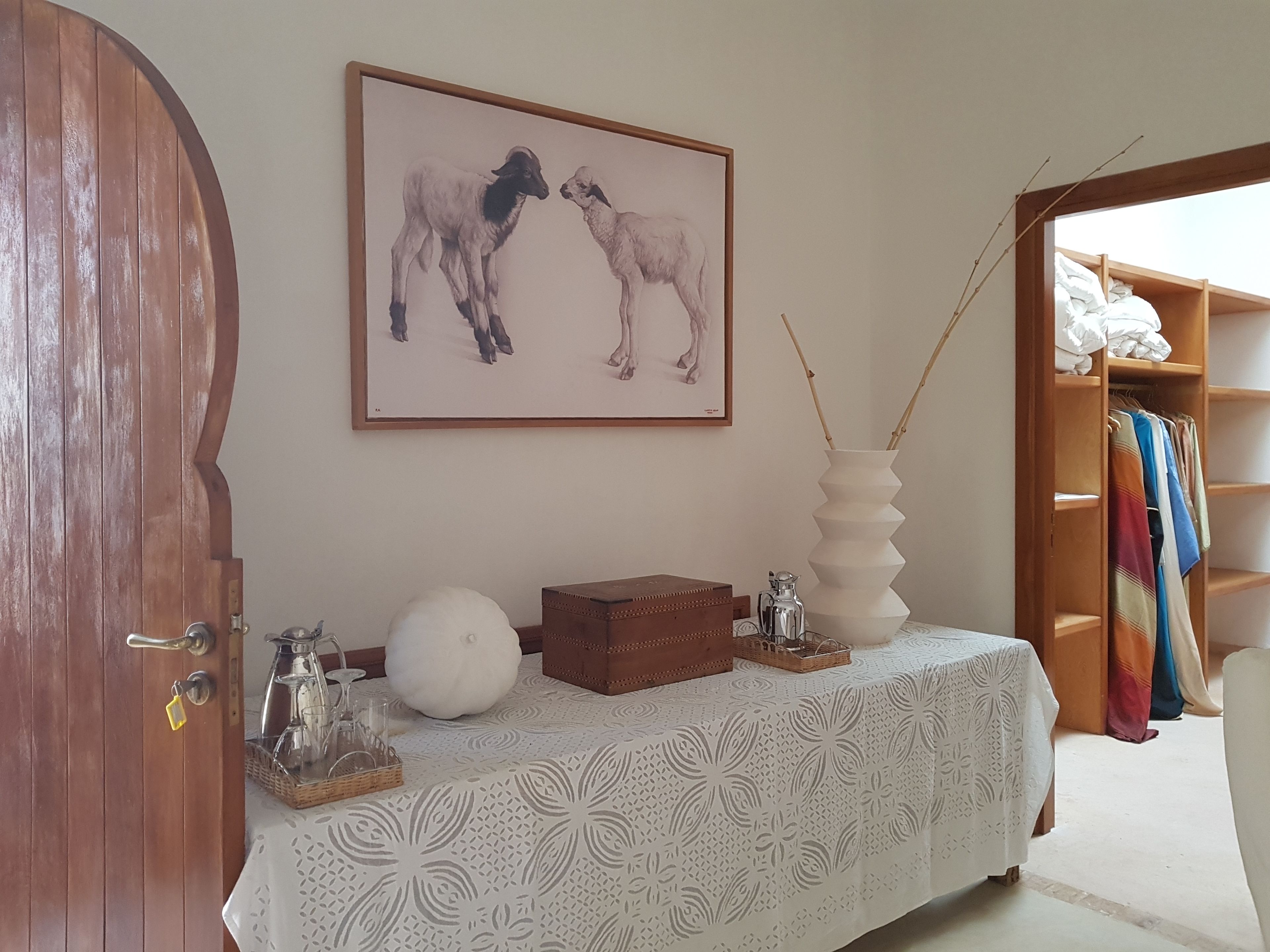 A room with a wooden door, a framed picture of two goats, a coffee maker, a white vase, and a wooden shelf with colorful clothes.