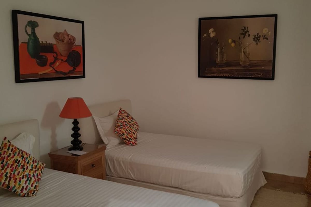 A bedroom with two single beds, a nightstand with a lamp, and two framed artworks on the wall.