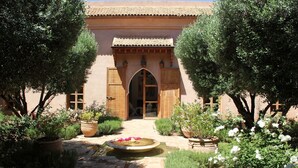 Un patio con una fuente, plantas en macetas y un edificio con tejado de azulejos y puertas de madera.