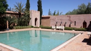 Una piscina rectangular con borde de azulejos, rodeada por un patio de ladrillo. Se ven palmeras y un muro con aberturas arqueadas.
