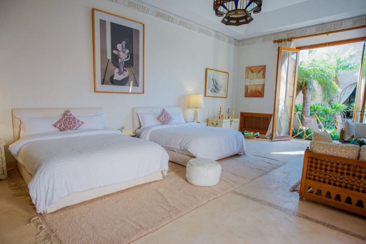 A bedroom with two beds, a small ottoman, and a wicker chair.