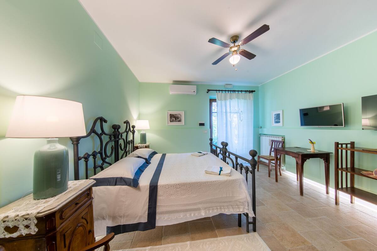 A bedroom with a bed, bedside tables, a desk, a chair, a ceiling fan, and a television.