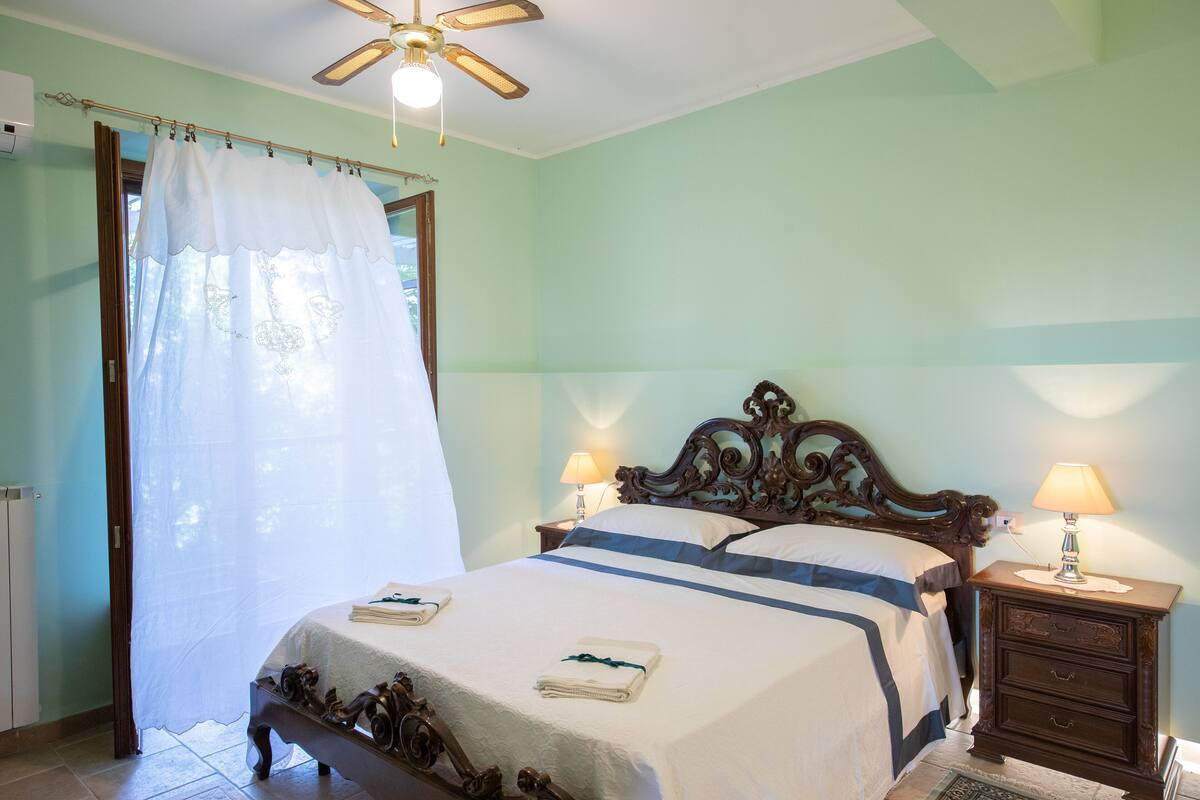 A bedroom with a bed, a nightstand with a lamp, a ceiling fan, and a window with a curtain.