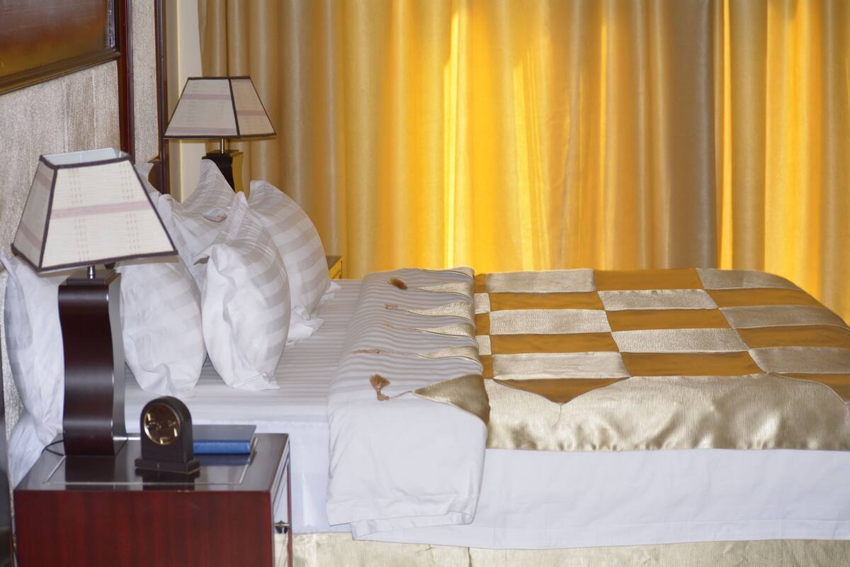 A hotel room with a neatly made bed, two bedside lamps, a wooden nightstand, and a large window with yellow curtains.