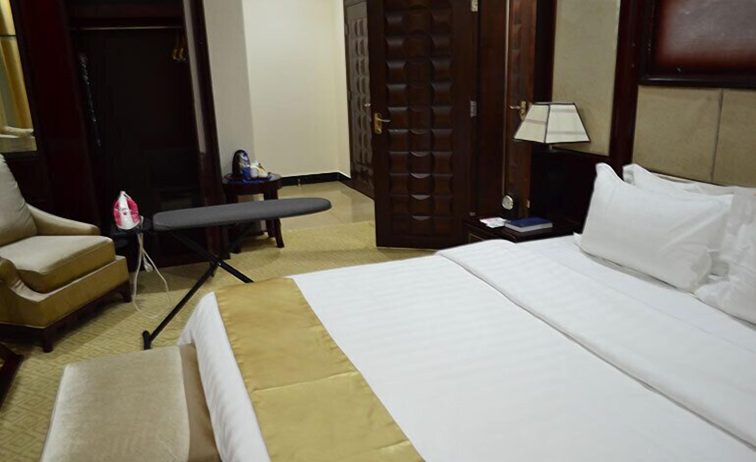 Una habitación de hotel con una cama, una tabla de planchar, una silla, una mesita de noche con lámpara y una puerta.