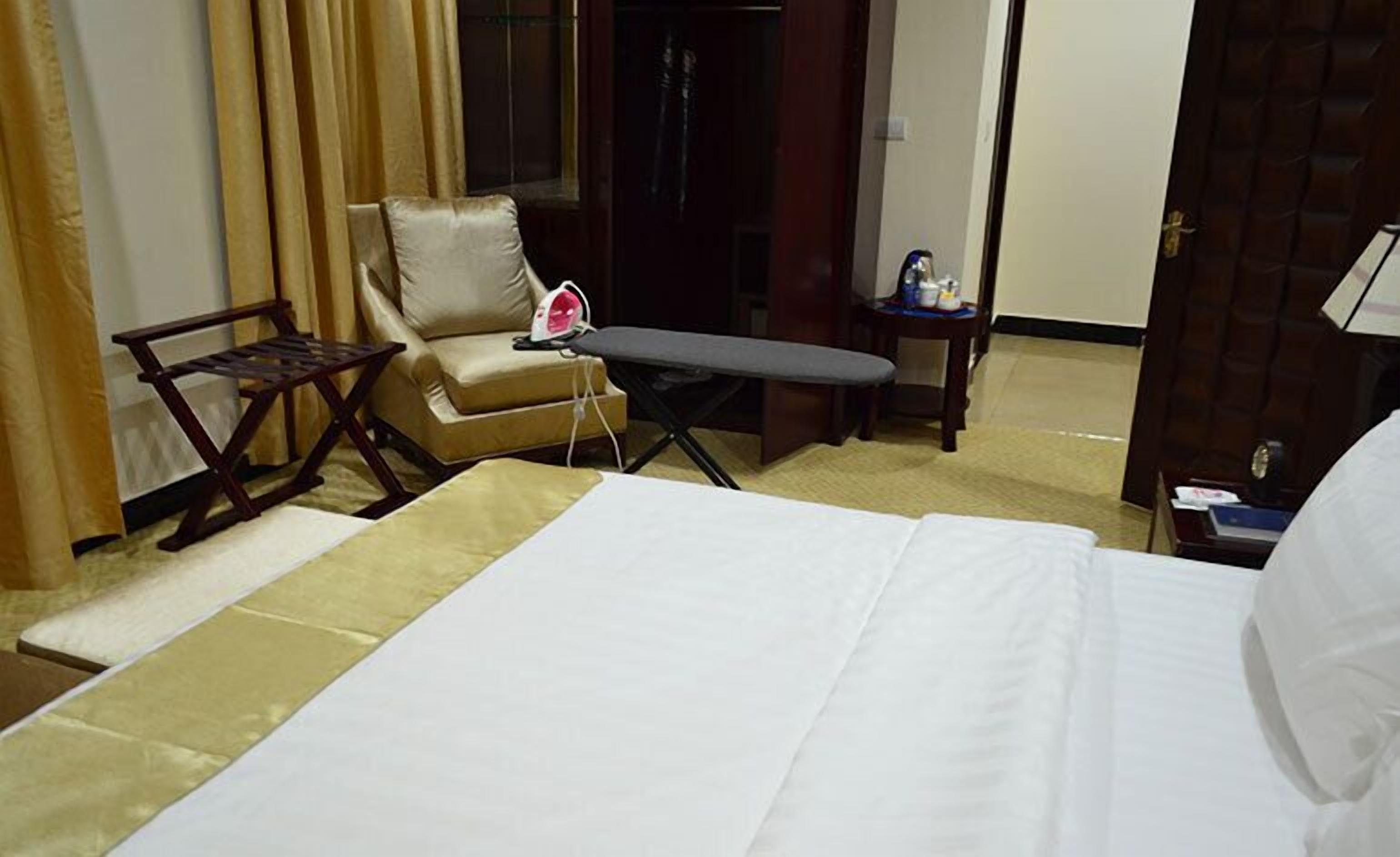 A hotel room with a bed, an ironing board, a chair, and a small table with a kettle.