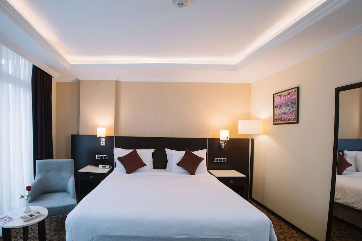 A hotel room with a large bed, two bedside tables with lamps, a small round table with a vase and a book, a chair, and a painting on the wall.
