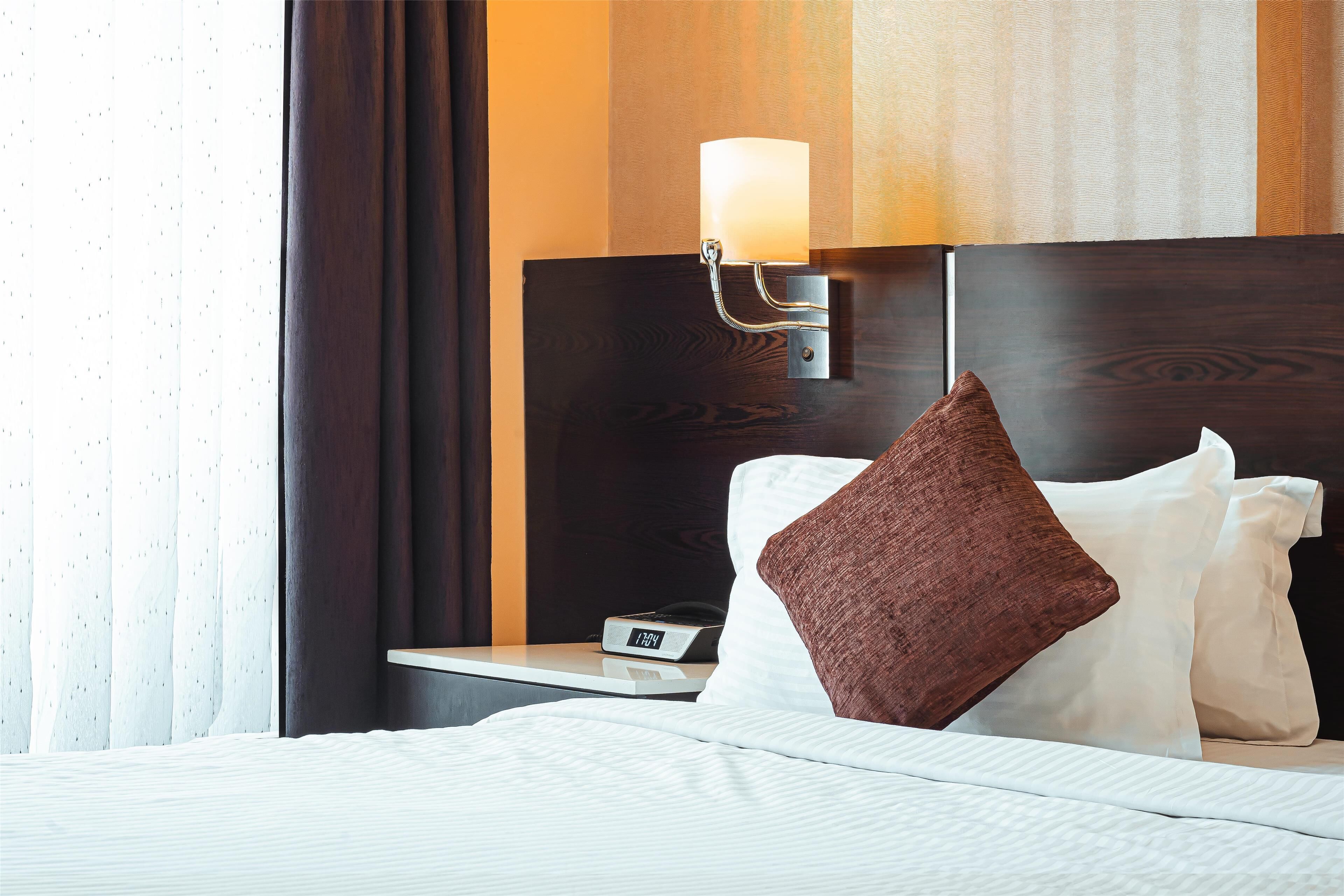 Un letto rifatto con lenzuola bianche, un cuscino marrone, un comodino con una lampada e una sveglia digitale che segna le 12:04.