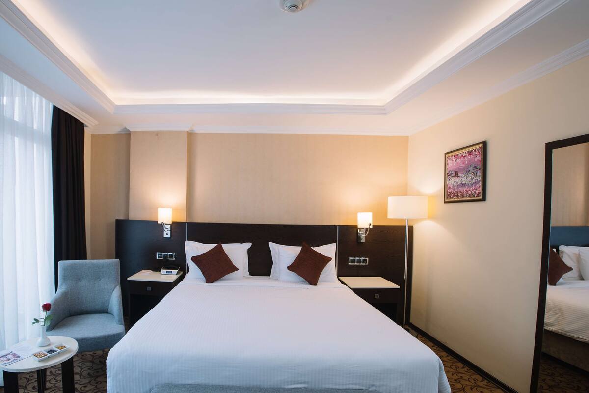 A hotel room with a large bed, two bedside tables, a chair, a small table with a vase and a book, and a painting on the wall.
