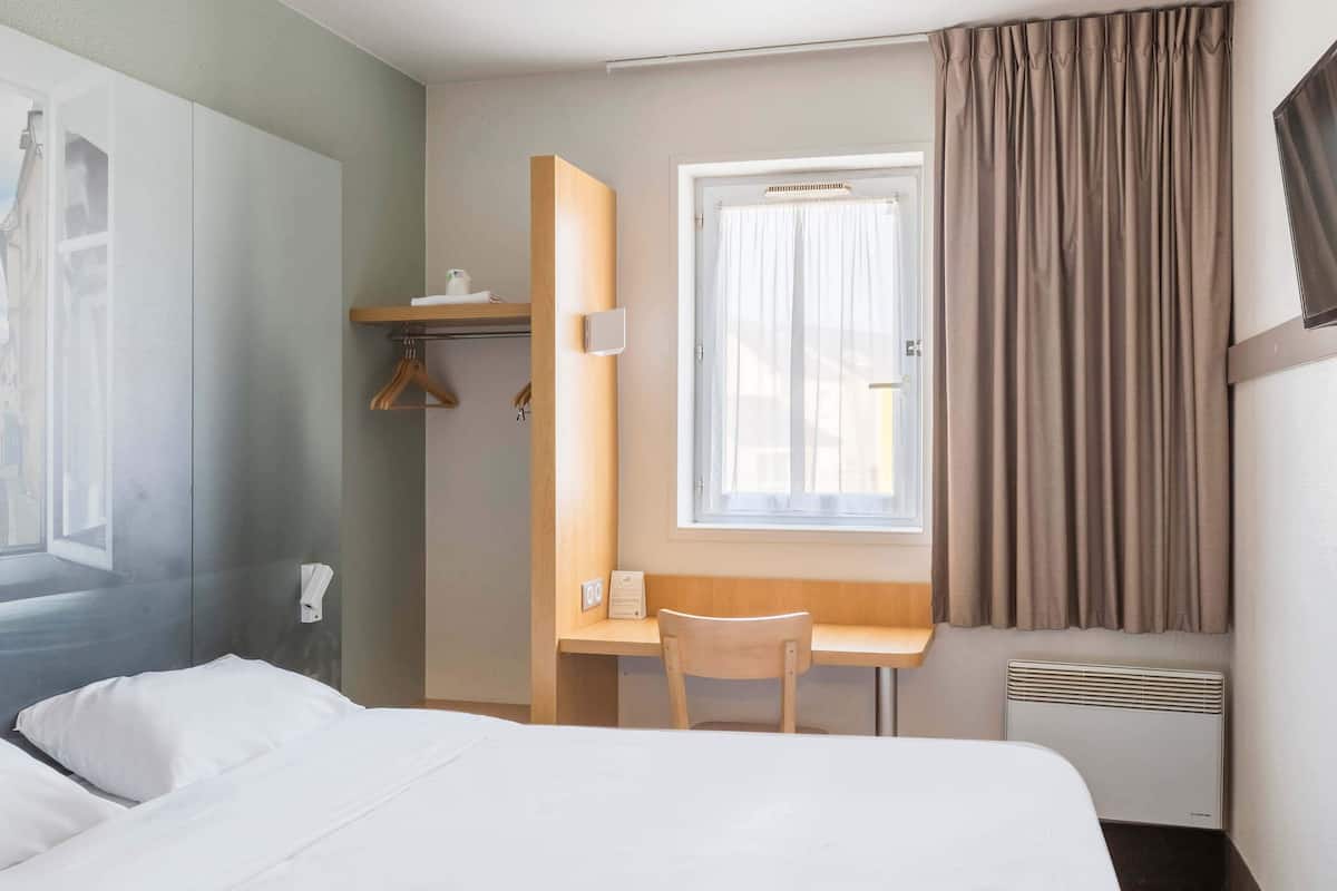 A hotel room with a bed, a desk, a chair, a window with curtains, a radiator, and a wall-mounted TV.