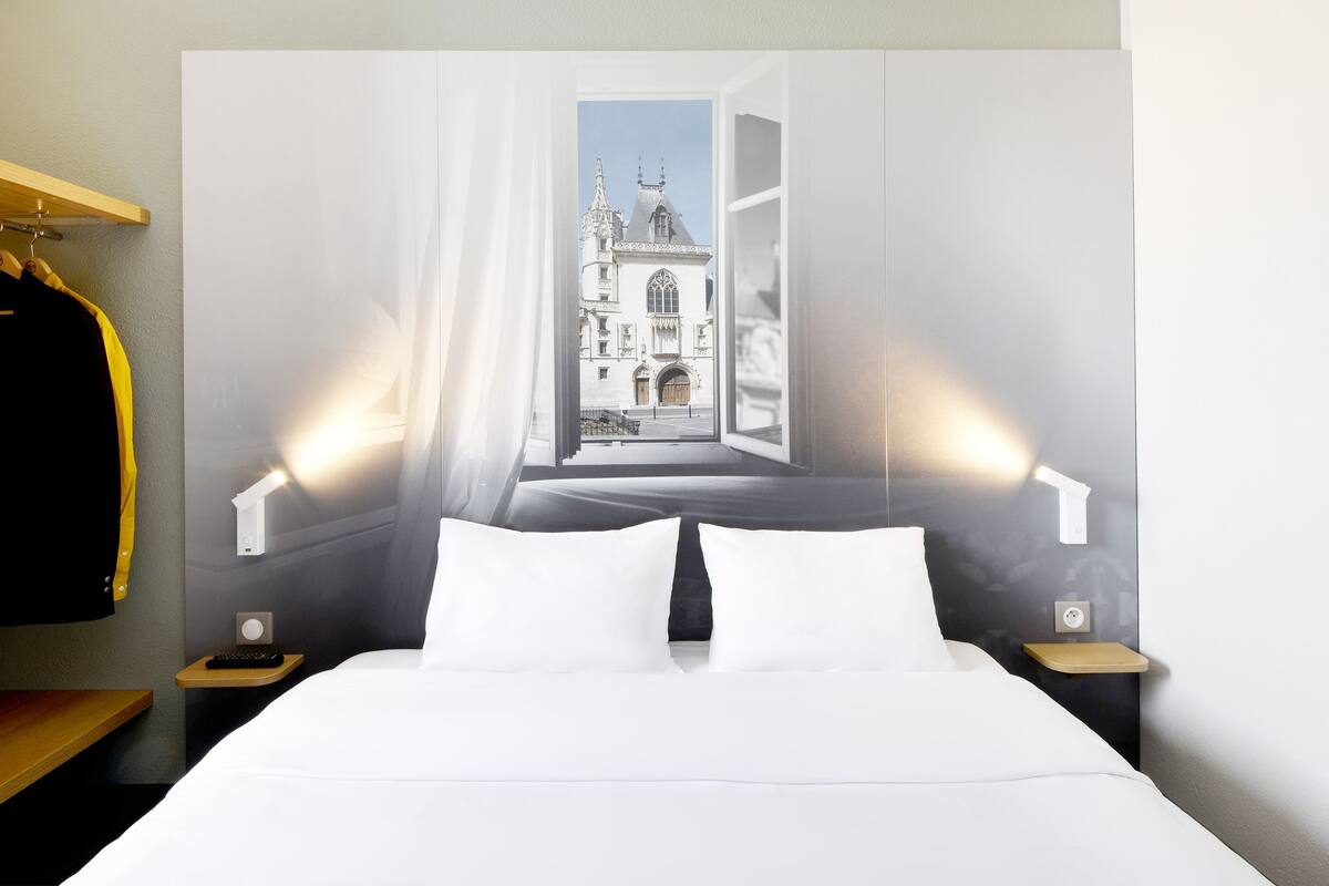 A hotel room with a bed, white pillows, a nightstand with a remote control, a wall-mounted lamp, and a view of a building through an open window.