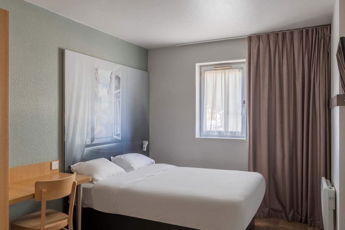 A neatly arranged hotel room with a bed, a small desk, a chair, a window with curtains, and a wall-mounted telephone.