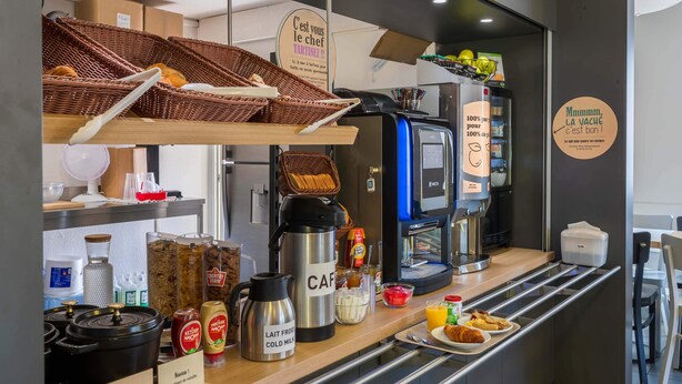 A cafe counter with a coffee machine, baskets of bread, condiments, and a sign reading "C'est vous le chef". - B&B HOTEL Bourges (1) (Bourges)
