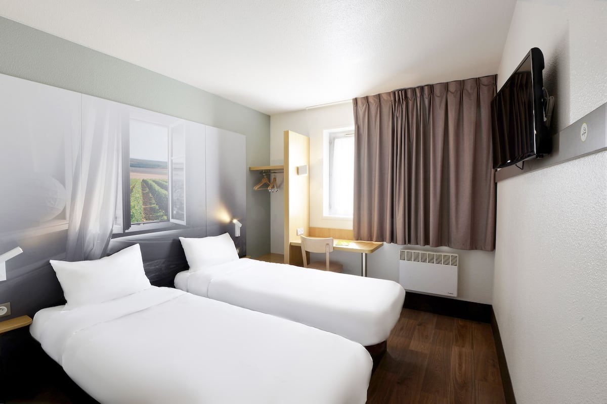 A hotel room with two beds, a desk, a chair, a TV, and a window with curtains.