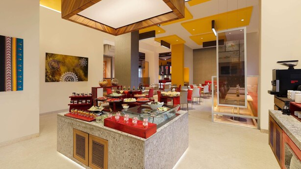 A modern hotel lobby with a central island displaying food and drinks, a coffee machine, and a seating area with tables and chairs. - ibis Pune Hinjewadi Hotel (Paud)