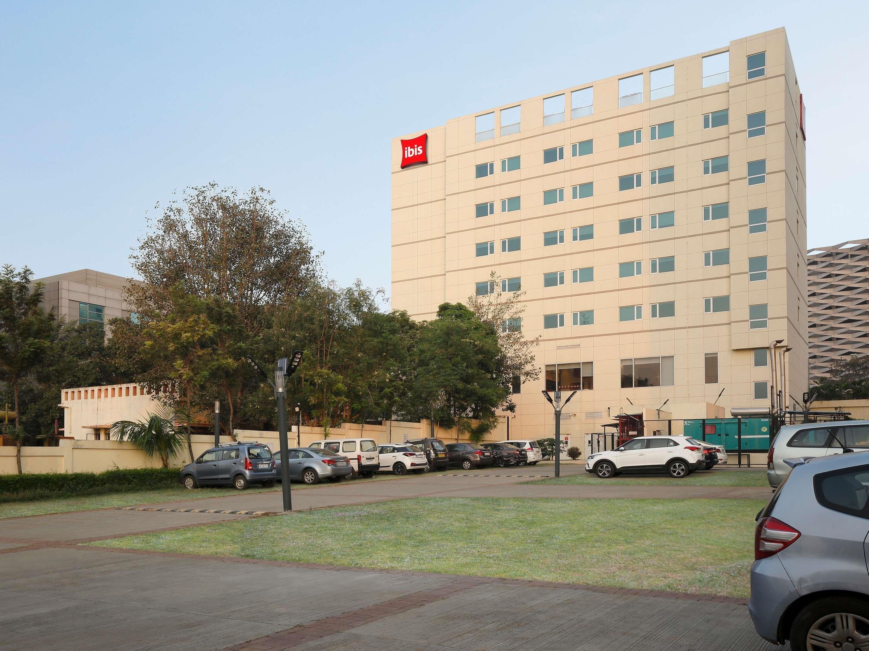 A multi-story hotel with a prominent 'ibis' sign, a parking lot with several cars, and a grassy area in front.
