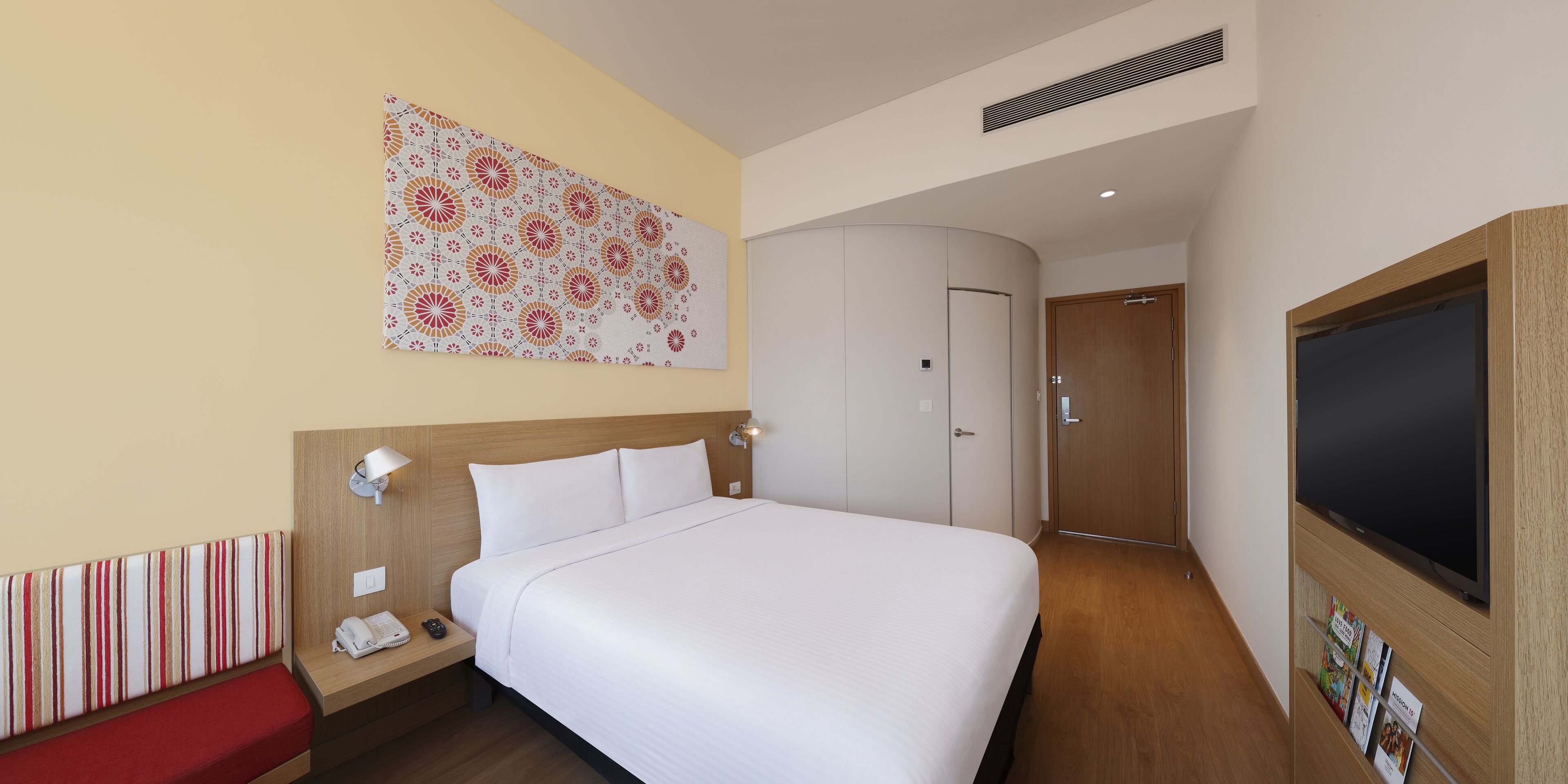 Habitación de hotel con una cama grande, una mesita de noche con lámpara, un banco, un armario y un televisor.