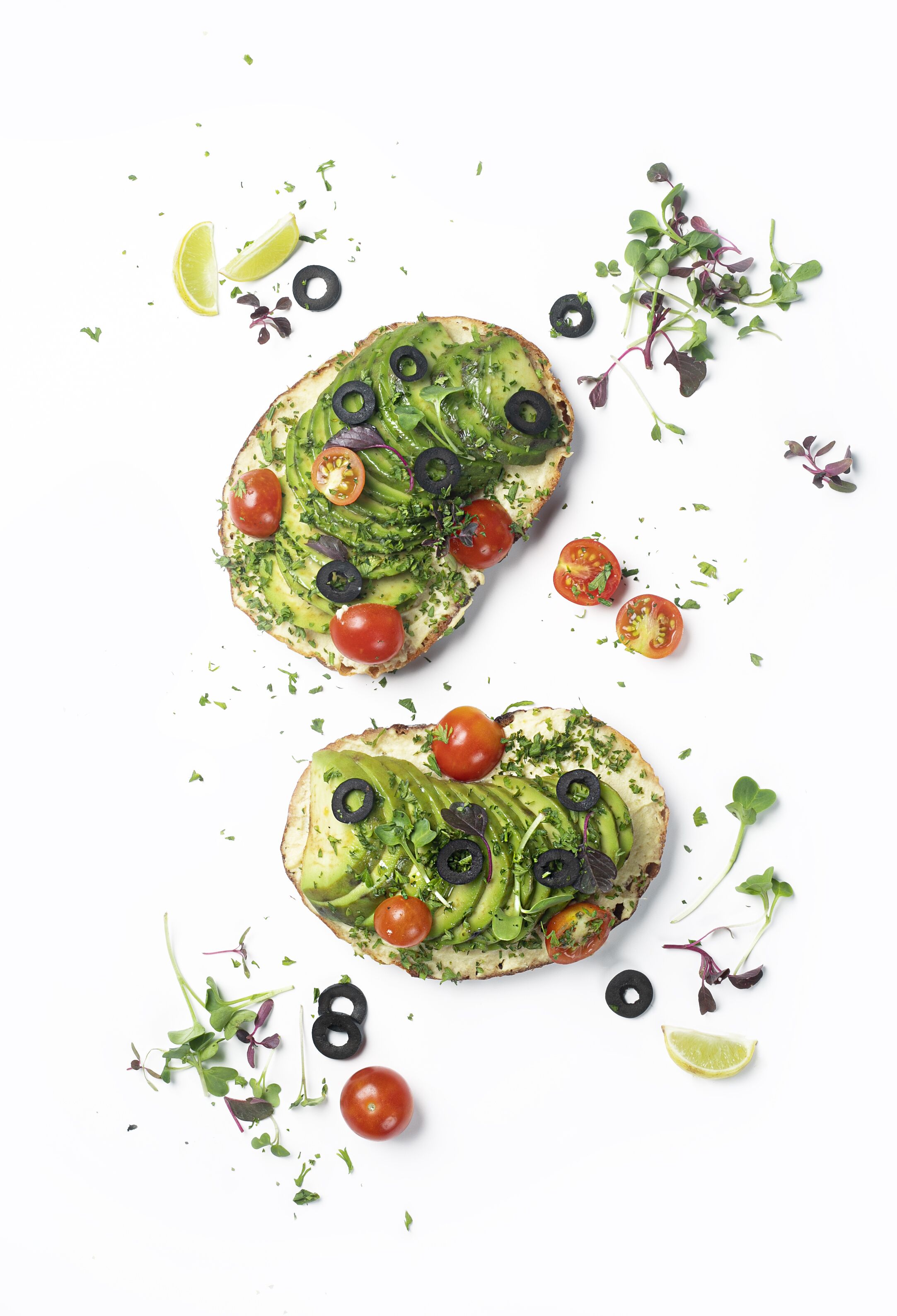 Deux sandwichs ouverts garnis d’avocat, de tomates et d’olives noires, présentés sur un fond blanc.