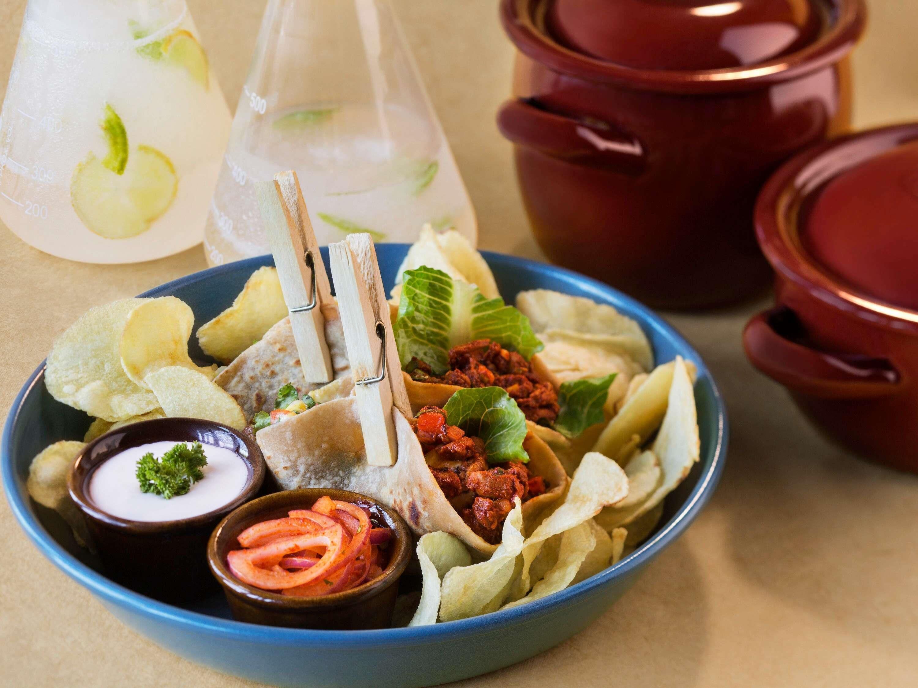 A bowl of food with chips, a dipping sauce, and a drink in the background.