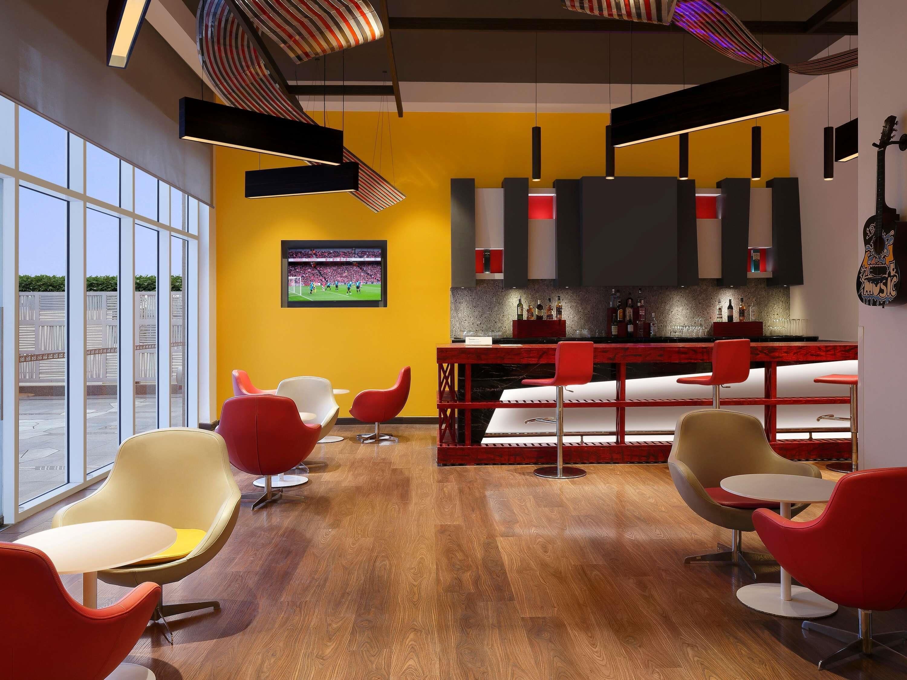 Un área de bar moderna con taburetes rojos, un televisor mostrando un evento deportivo y un amplio ventanal con vistas al exterior.