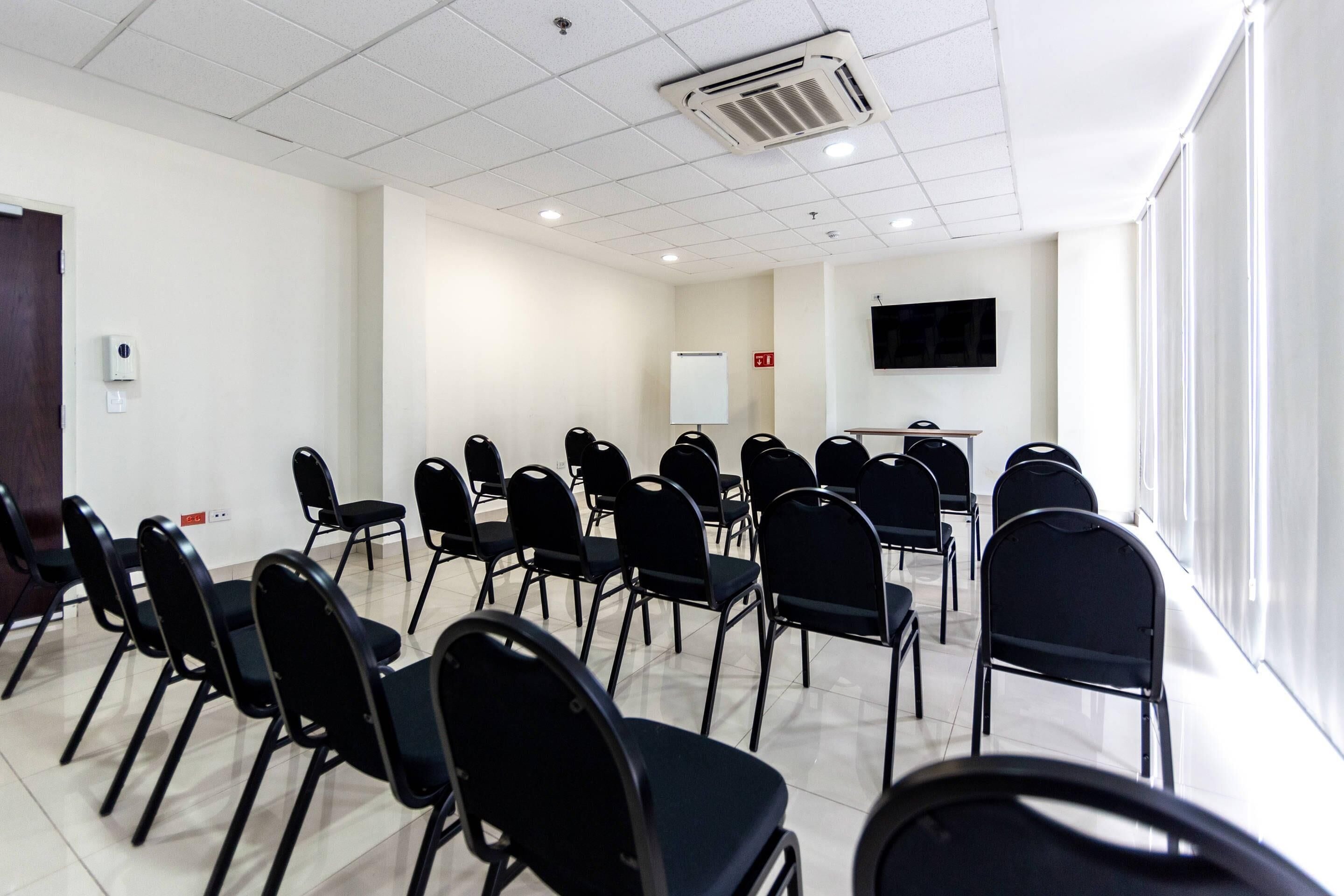 A conference room with rows of black chairs, a whiteboard, a flat-screen TV, and an air conditioning unit on the ceiling.