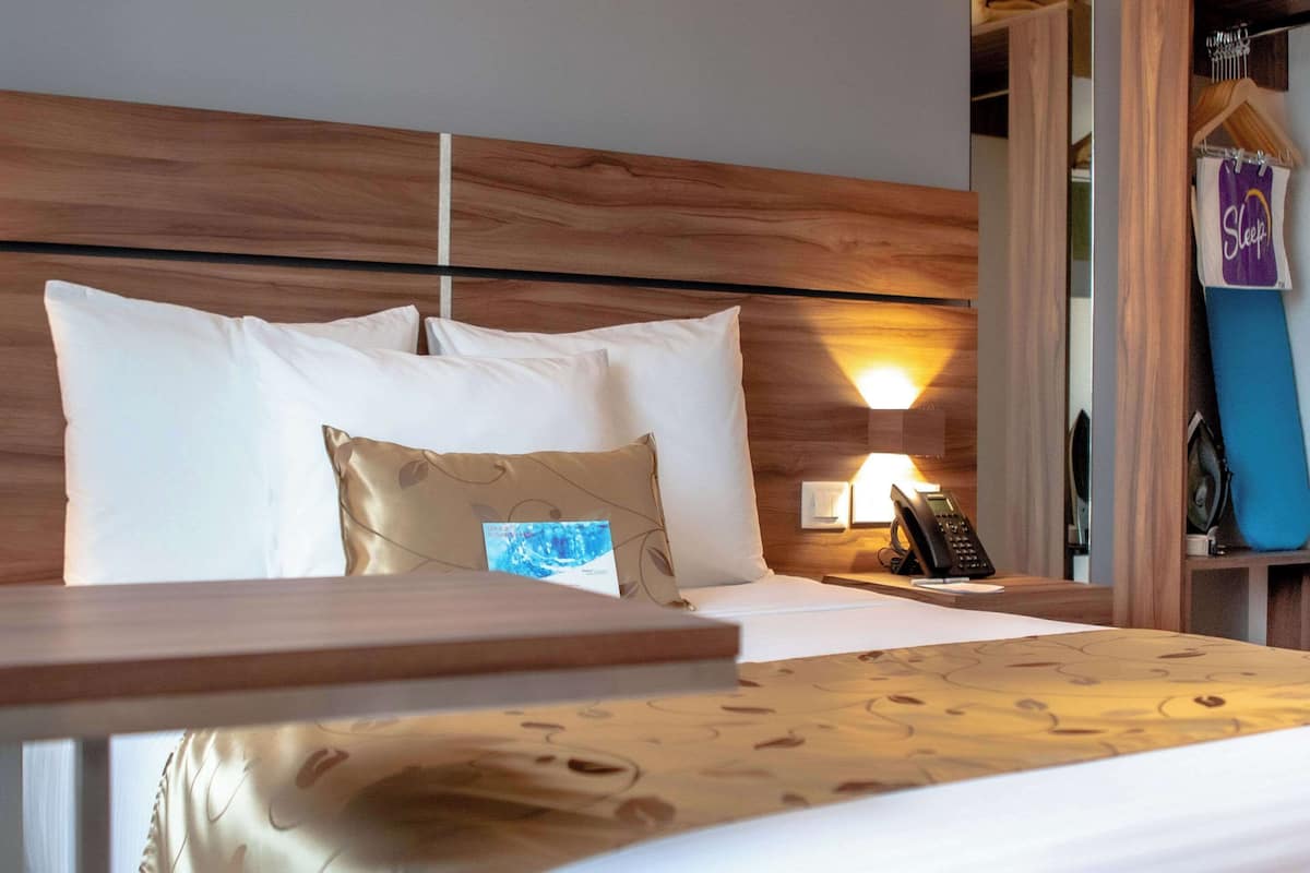 Une chambre d’hôtel avec une tête de lit en bois, un lit avec des oreillers blancs et à motifs, une table de chevet, une lampe et une armoire où est suspendu un vêtement.