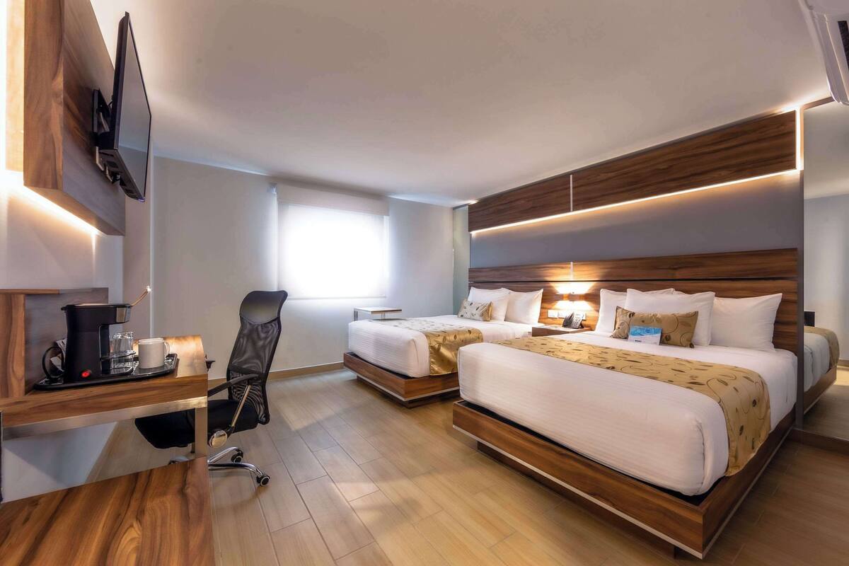 Une chambre d’hôtel moderne équipée de deux lits, d’un bureau avec une machine à café, d’une chaise et d’un téléviseur fixé au mur.