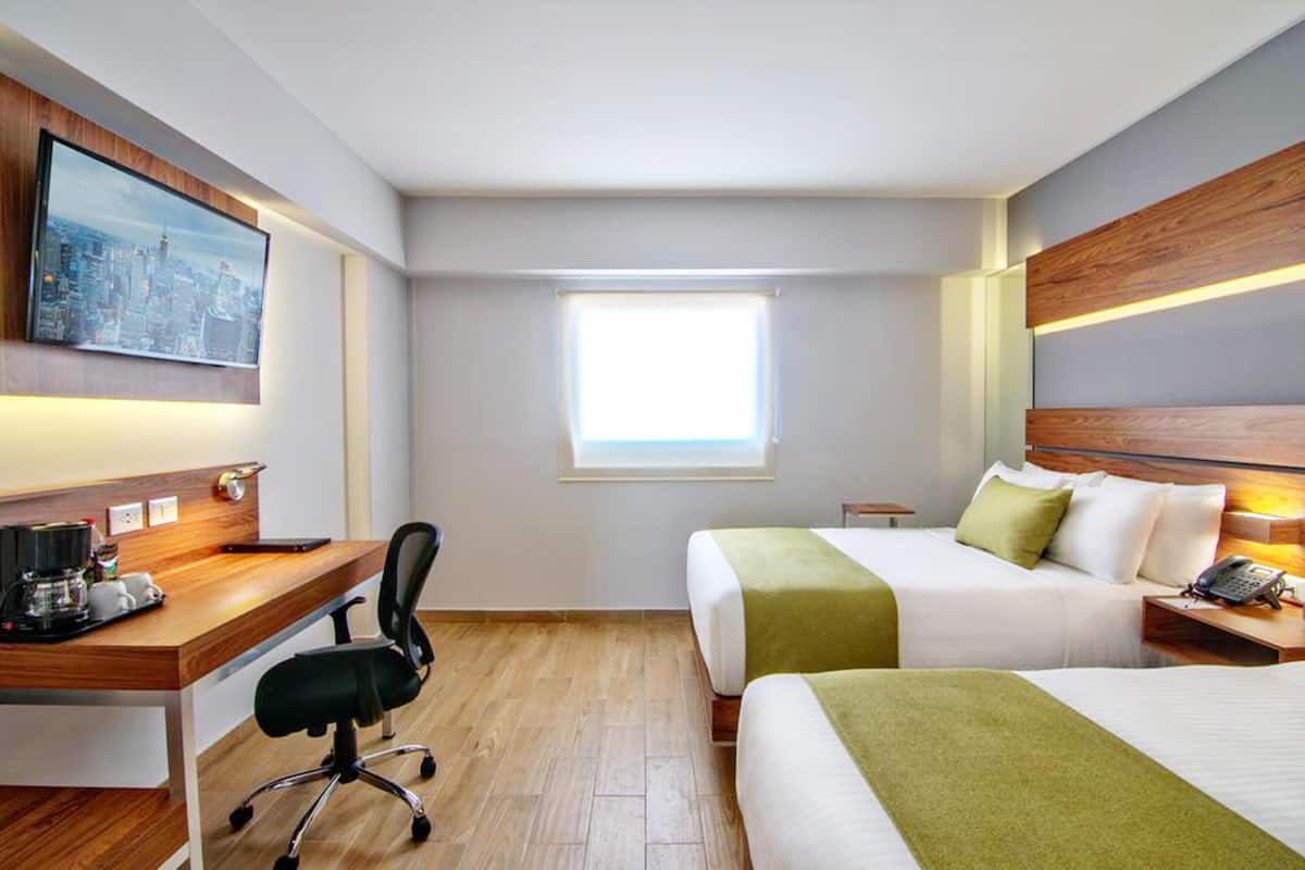 Une chambre d’hôtel avec un bureau, deux lits, une fenêtre et un tableau représentant un paysage urbain.