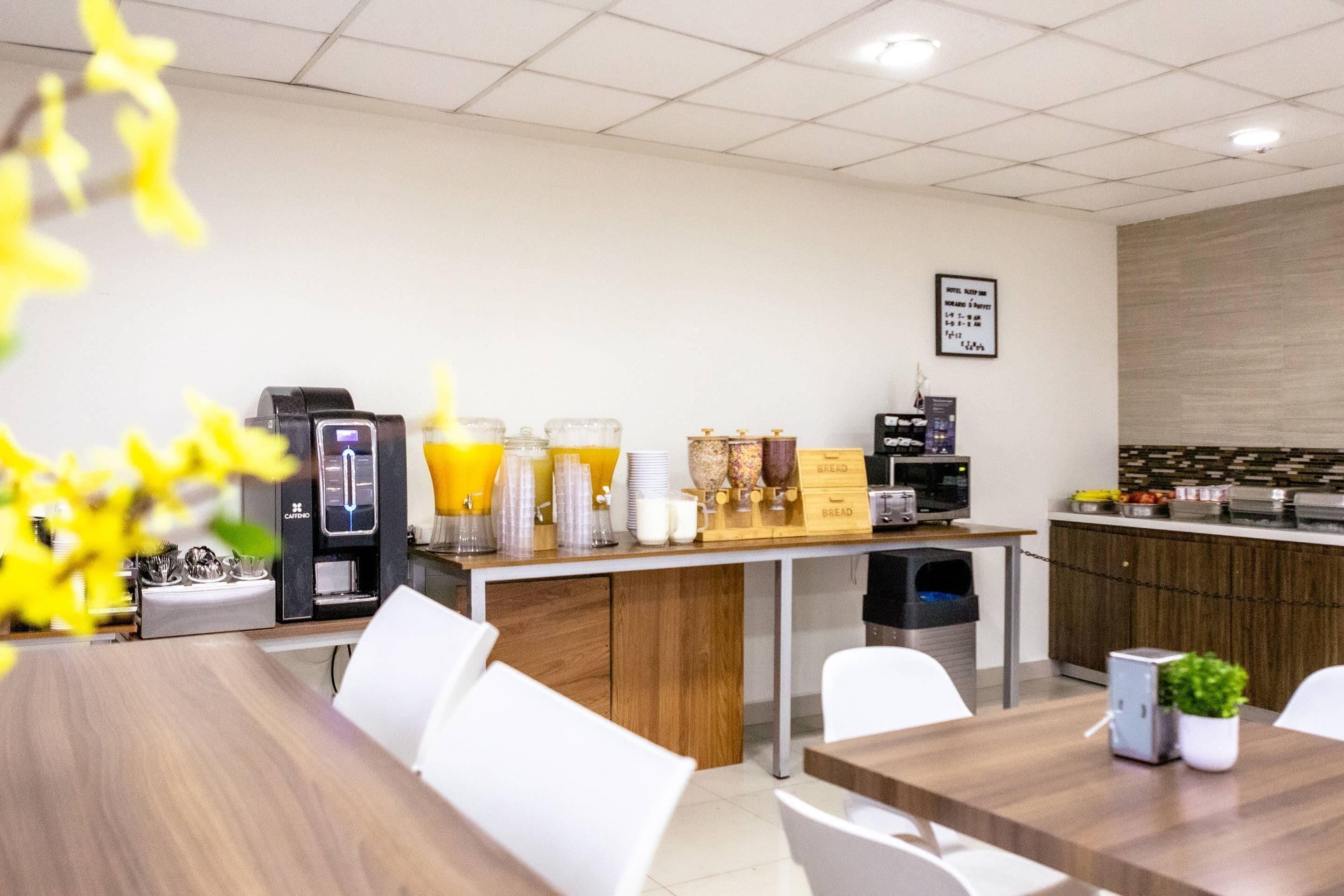 A modern kitchen with a coffee machine, a juice dispenser, and a microwave. There is a wooden counter with various food items and a sign indicating breakfast hours.