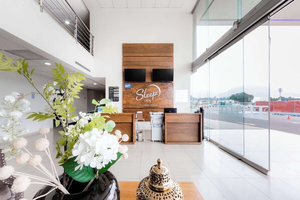 A hotel reception area with a wooden reception desk, a large window offering a view of the outside, and a decorative plant with white flowers. - Sleep Inn Tijuana (Tijuana)