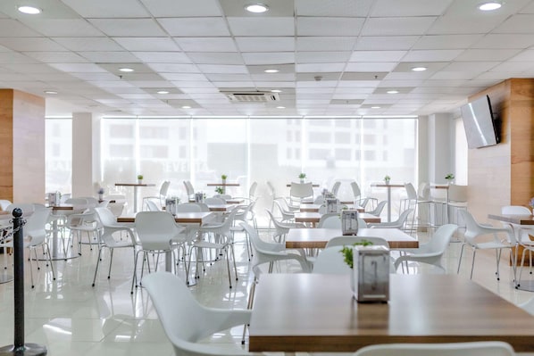 Un comedor amplio y moderno con sillas y mesas blancas, ventanas grandes y un televisor de pantalla plana montado en la pared.