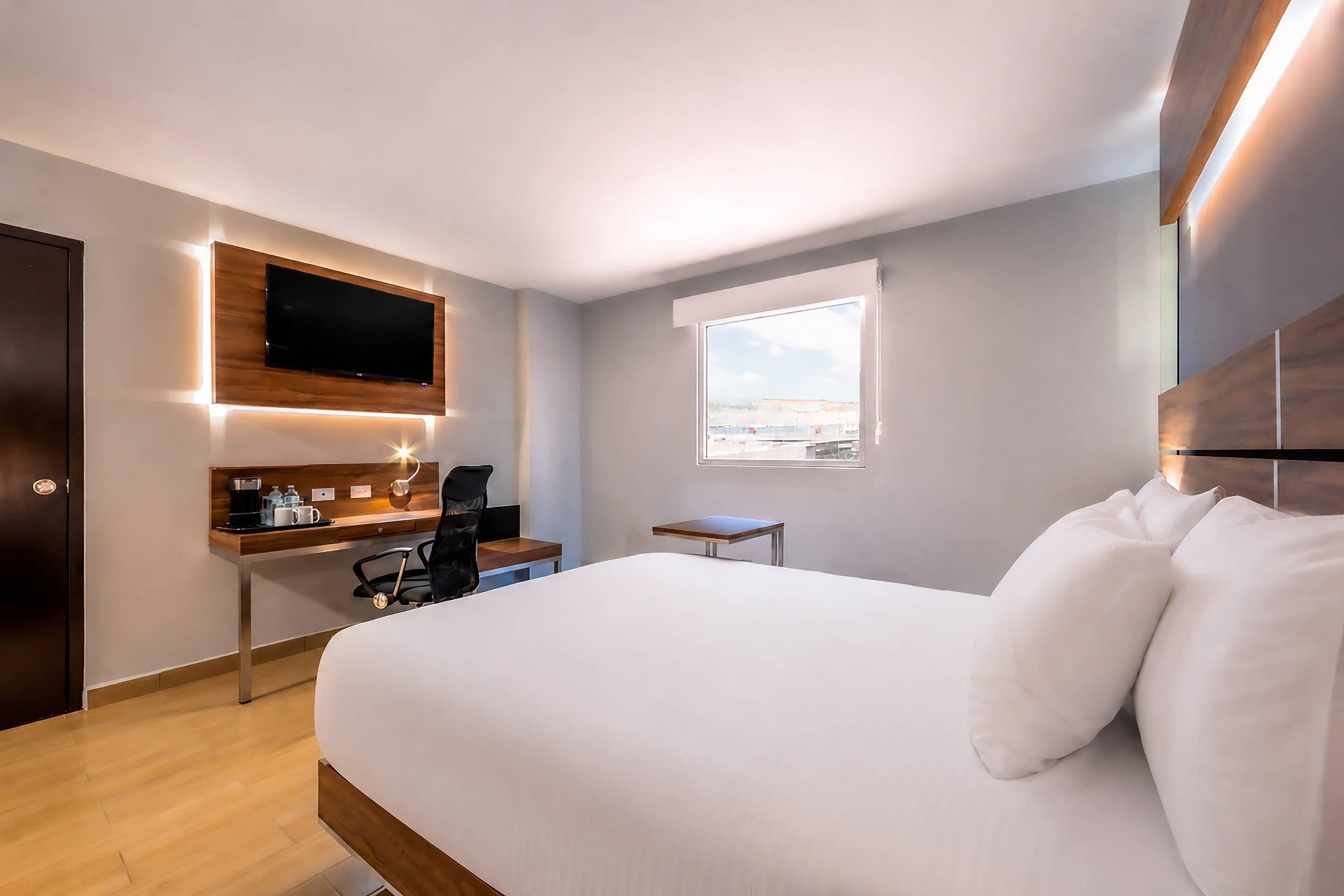 A hotel room with a bed, a desk with a chair, a television mounted on the wall, and a window providing a view.