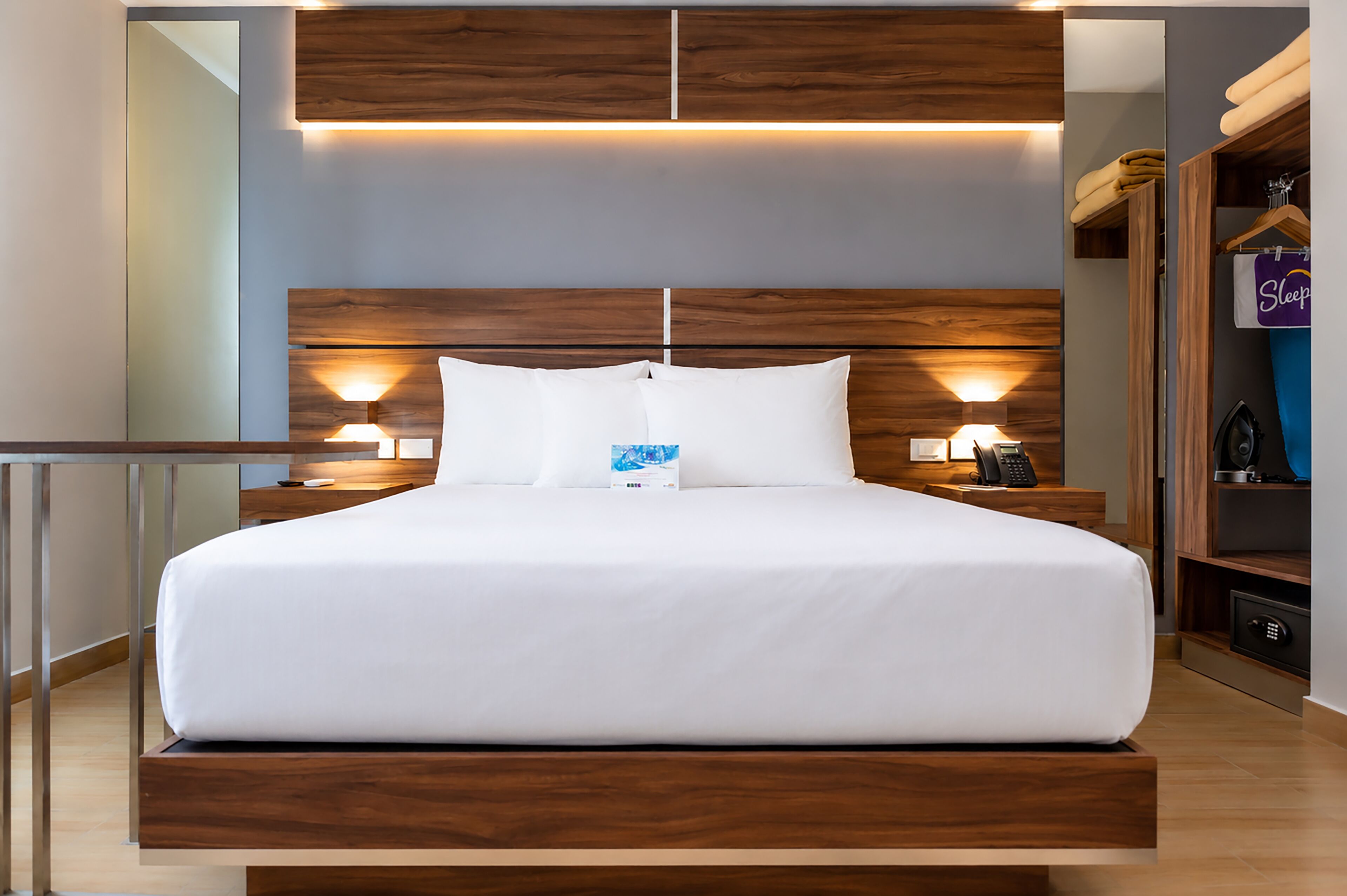 A modern hotel room with a large bed, wooden headboard, bedside lamps, a bedside table, and a wardrobe with a hanging bag.