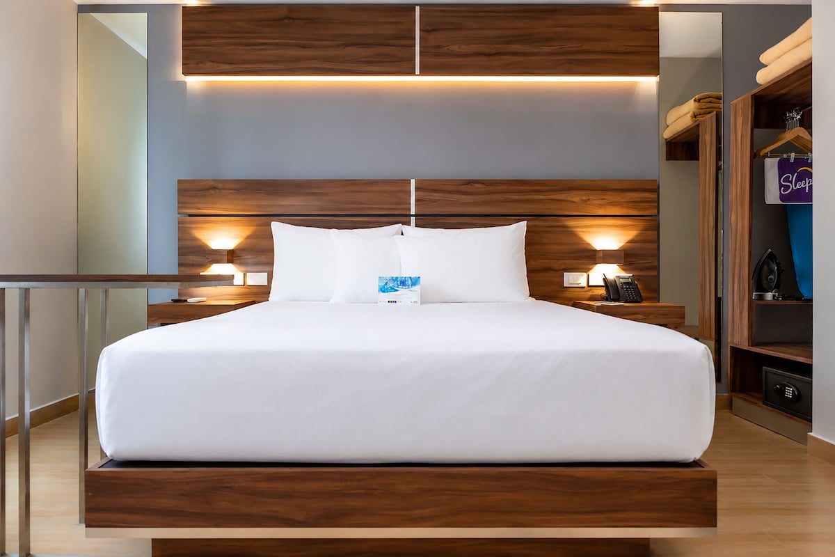 A modern hotel room with a large bed, wooden headboard, bedside lamps, a bedside table, and a wardrobe with a hanging bag.