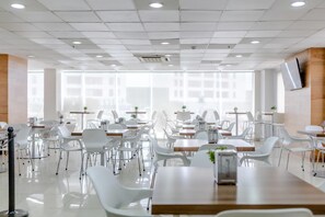 A modern dining area with white tables and chairs, large windows, and a flat-screen TV mounted on the wall. - Sleep Inn Tijuana (Tijuana)
