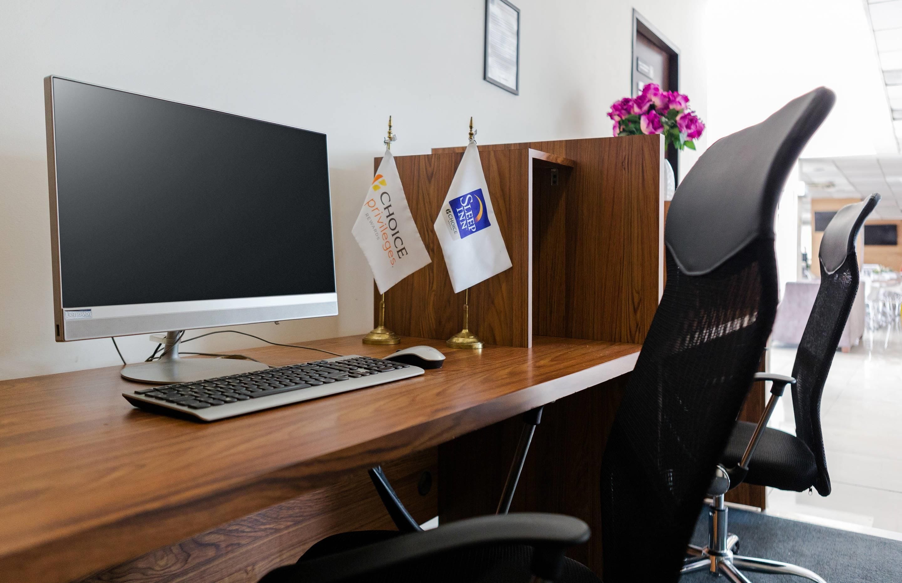 Una moderna scrivania da ufficio con un computer, una tastiera, un mouse e una sedia da ufficio nera. Sulla parete sono appese due bandiere, una con il logo 'Choice Privileges' e l'altra con 'Sleep Inn'.