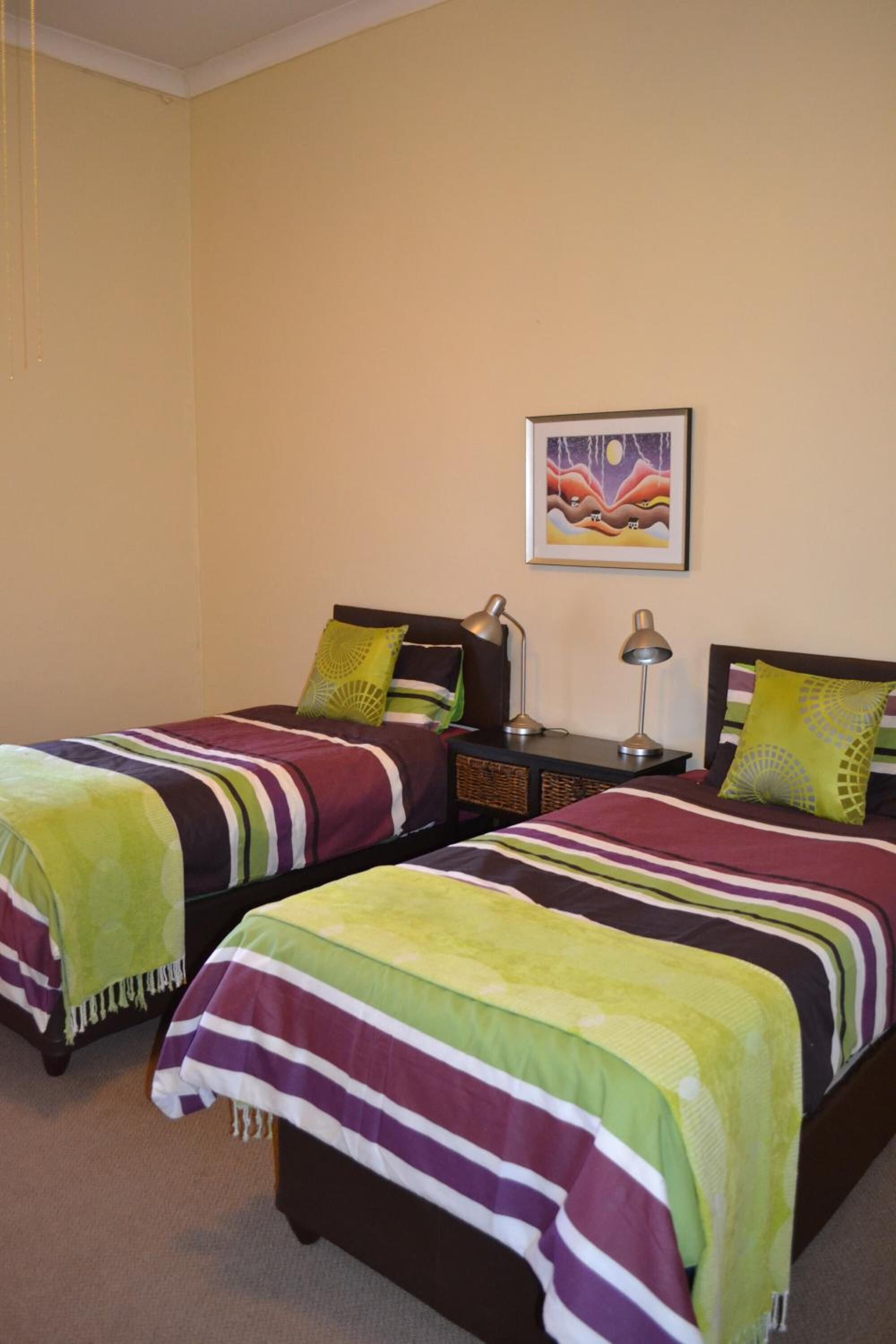 A hotel room with two beds, each with a striped bedspread and a green throw blanket. There is a nightstand with a lamp and a framed picture on the wall.