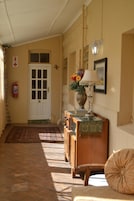Un pasillo con piso de baldosas, una puerta blanca, un extintor, un banco de madera, una mesa con una lámpara y flores, y un aplique en la pared.