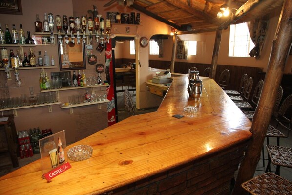 A bar area with a long wooden counter, shelves with various bottles and glasses, and wicker chairs around the bar.