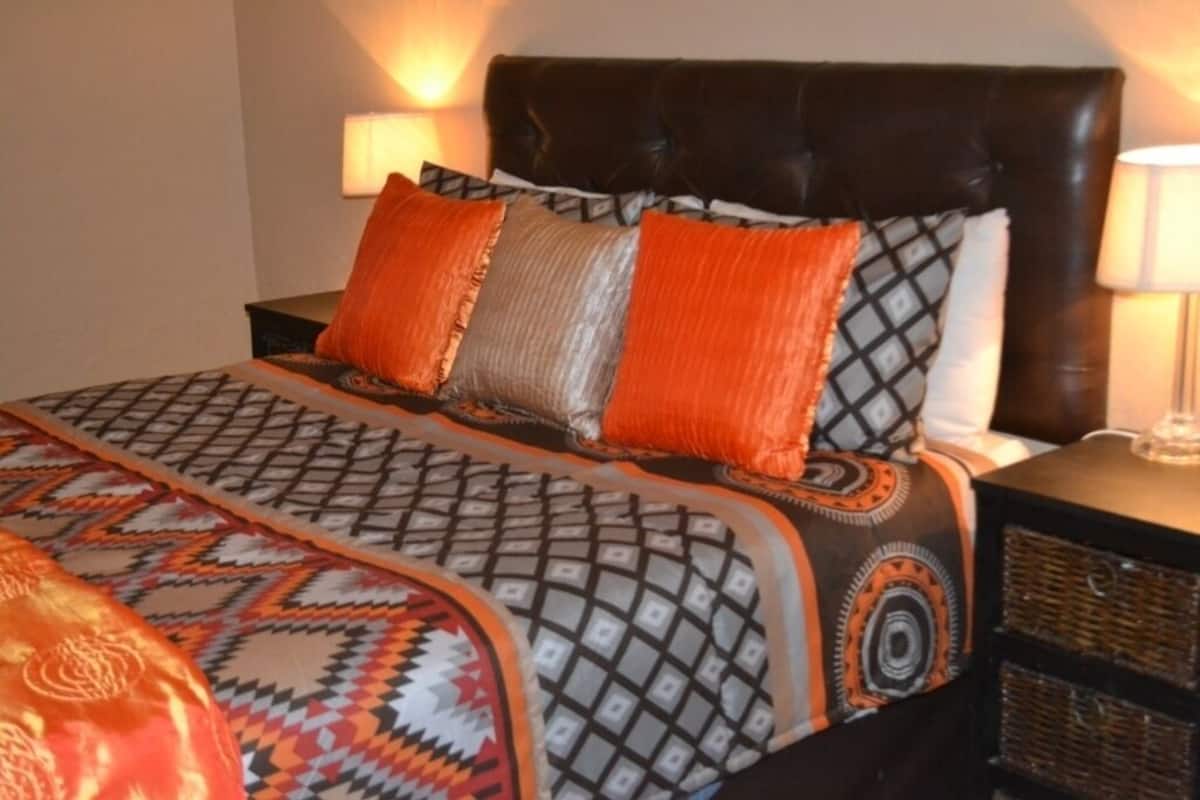 Ein Bett mit einem gemusterten Überwurf und zwei Kissen, ein dunkles Kopfteil sowie ein Nachttisch mit einer Lampe und einem Korb.