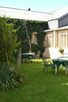 Un jardín con una mesa y sillas, rodeado de vegetación y un edificio con techo metálico.