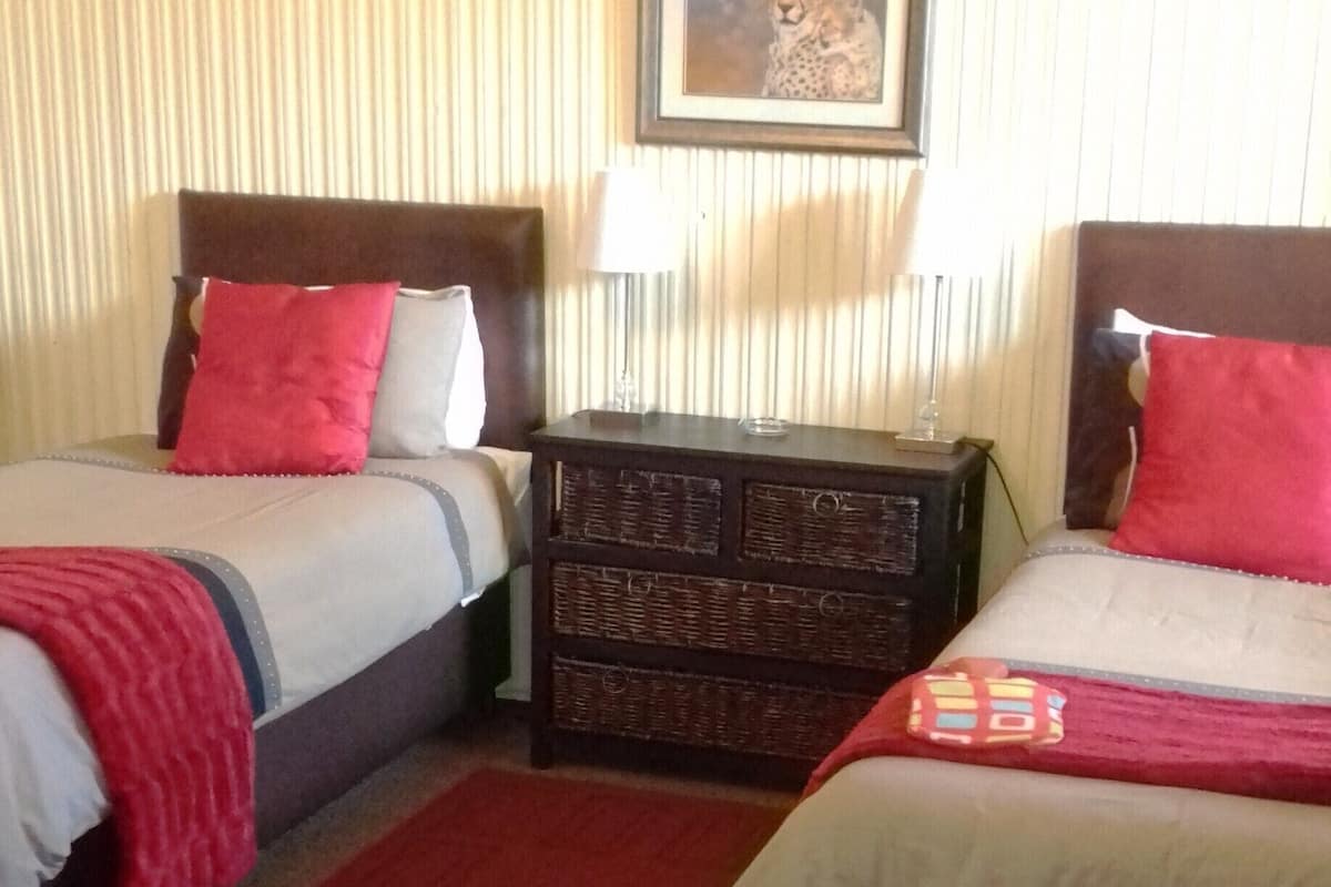 Ein Hotelzimmer mit zwei Betten, jedes mit einem roten Kopfkissen und einer weißen Bettdecke. Es gibt einen kleinen Nachttisch mit einer Lampe und ein gerahmtes Bild an der Wand über den Betten.